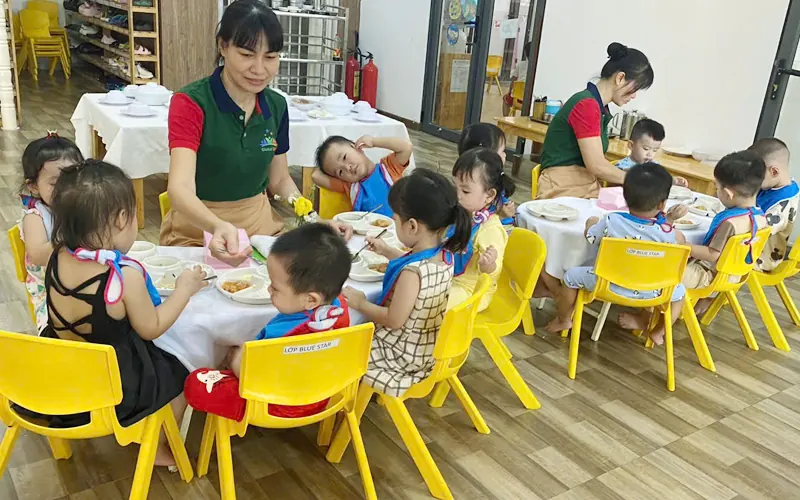 A teacher kneeling beside three young children who hold magnifying glasses over fallen leaves on a shaded preschool path, seen from behind, soft morning light
