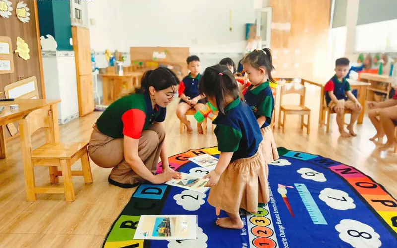 A Vietnamese teacher and an international teacher kneeling with four young children around a low table of leaves and wooden bowls, morning light, seen from behind
