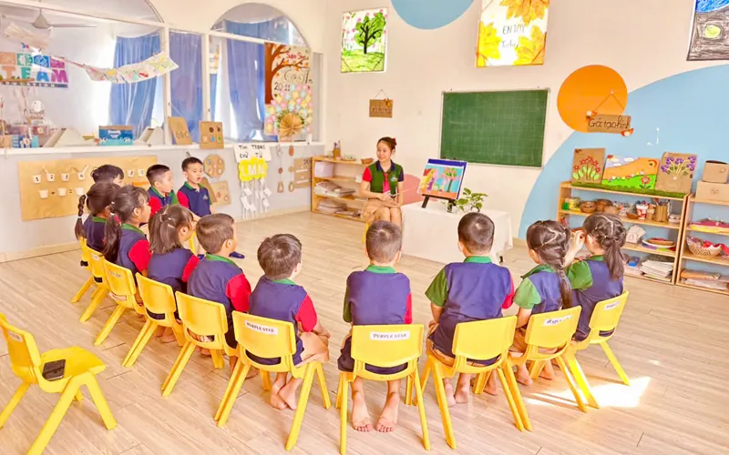 A Vietnamese teacher and an international teacher sitting together at a low wooden table after class, a closed laptop and a stack of children's clay models between them, evening light